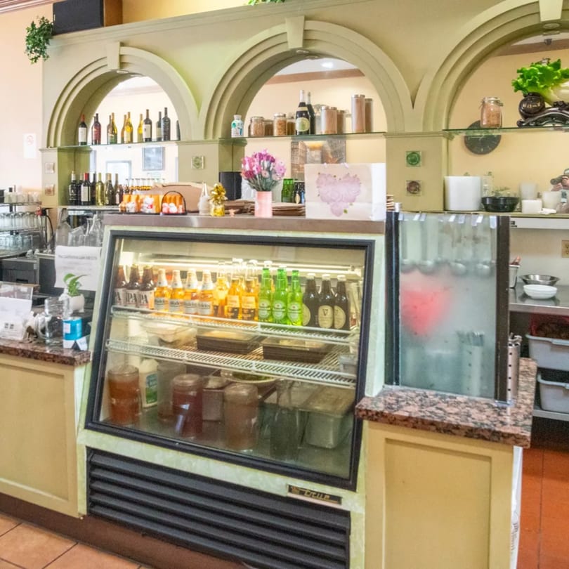 A beverage cooler filled with drinks, next to a counter with bottles and decor at Sage Bistro.