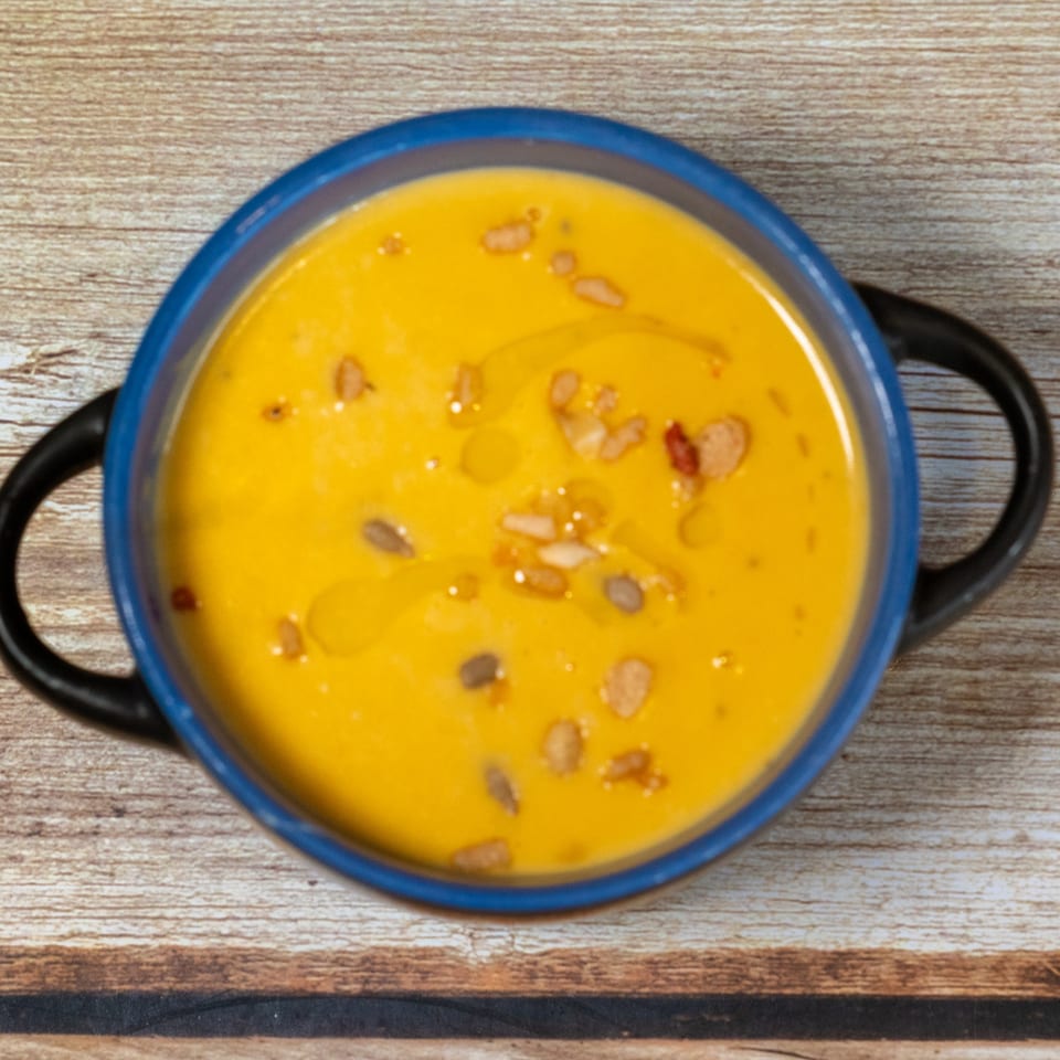 Overhead view of a vibrant orange Sage Bistro soup in a blue bowl with handles, garnished with seeds and oil.