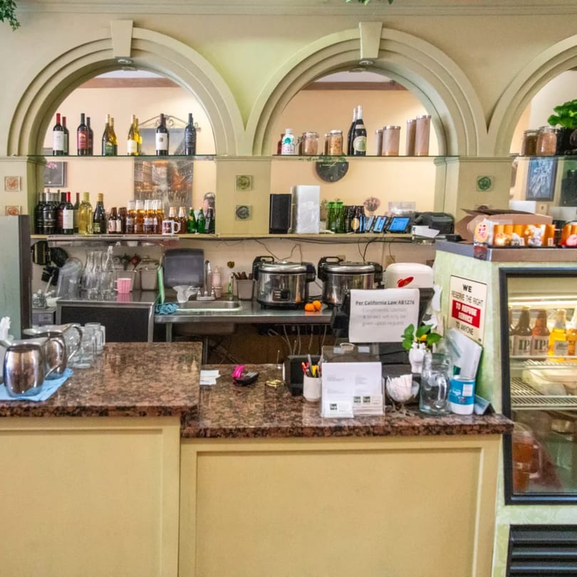 A Sage Bistro counter with bottles, kitchen equipment, and a refrigerated display case.