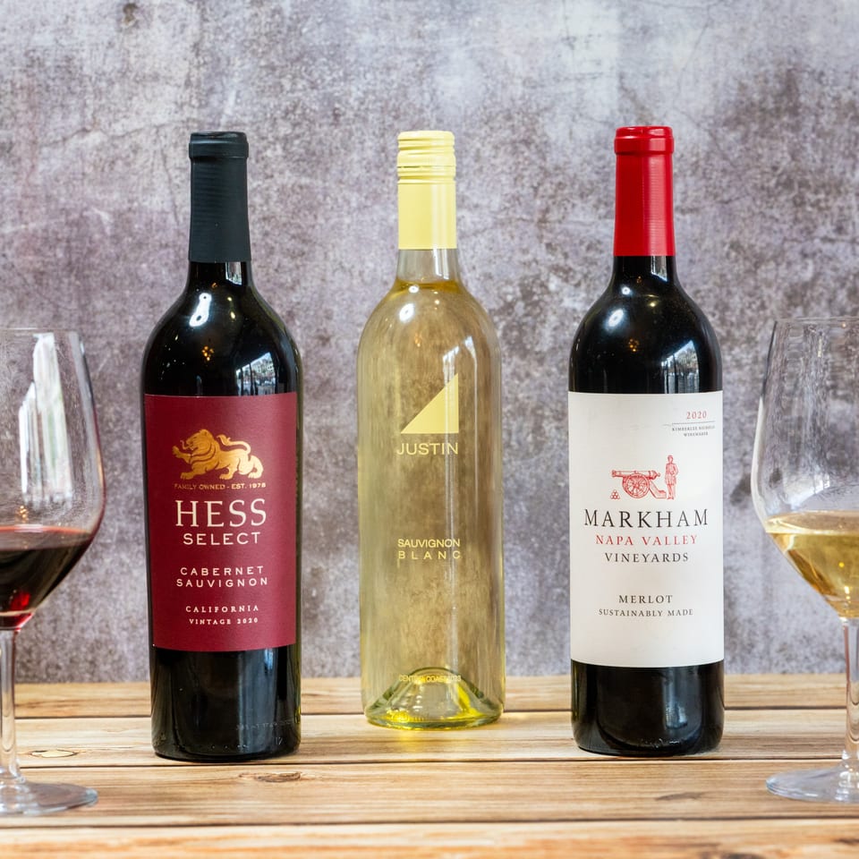 Three wine bottles, red and white, with two wine glasses on a rustic wooden table.